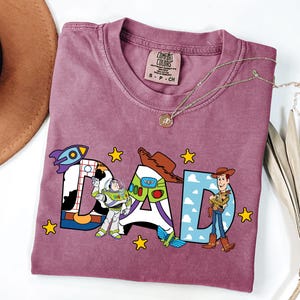 K&ouml;nnte beinhalten: Ein malvenfarbenes Kurzarm-T-Shirt mit einem Grafikdruck des Wortes DAD. Die Buchstaben zeigen Muster aus Kuhflecken, einem Weltraum-Ranger-Anzug und blauem Himmel mit Wolken. Die Figuren Buzz Lightyear und Woody sind neben den Buchstaben abgebildet.