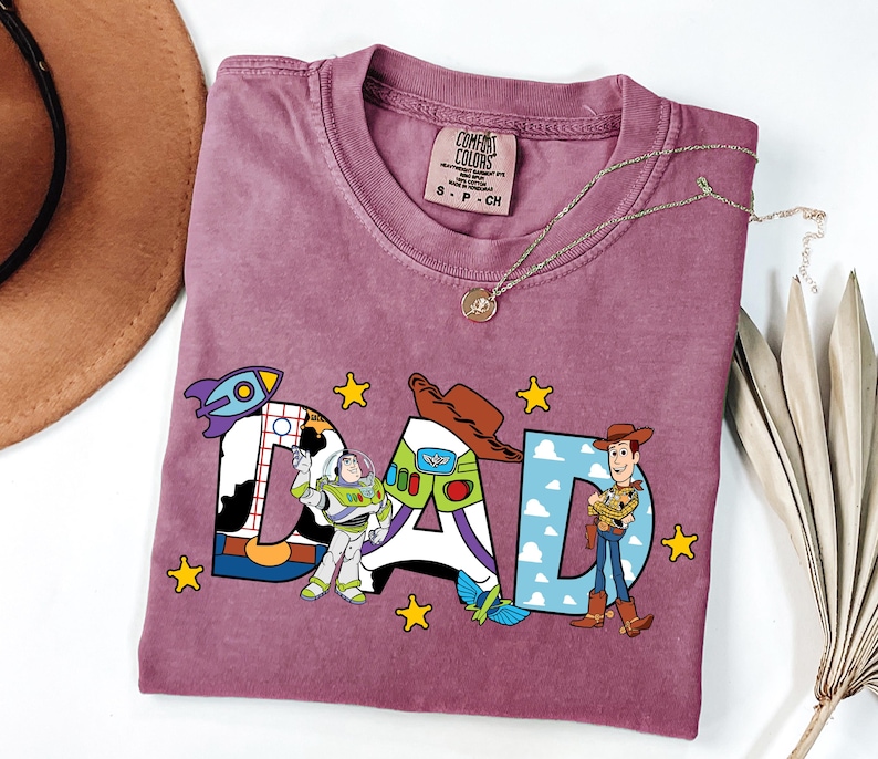K&ouml;nnte beinhalten: Ein malvenfarbenes Kurzarm-T-Shirt mit einem Grafikdruck des Wortes DAD. Die Buchstaben zeigen Muster aus Kuhflecken, einem Weltraum-Ranger-Anzug und blauem Himmel mit Wolken. Die Figuren Buzz Lightyear und Woody sind neben den Buchstaben abgebildet.