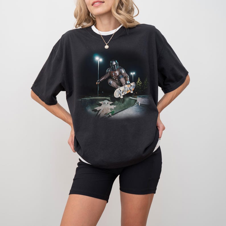 May include: Black t-shirt featuring a graphic of The Mandalorian skateboarding in a skate park with Grogu. The image is set at night with streetlights. The shirt has a white collar and is worn with black shorts.