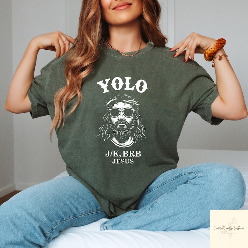Peut inclure: T-shirt vert olive avec le texte "YOLO" au-dessus d'un dessin de J&eacute;sus portant des lunettes de soleil et une couronne d'&eacute;pines. Le texte "J/K, BRB -JESUS" est en dessous. Le t-shirt est port&eacute; avec un jean bleu.