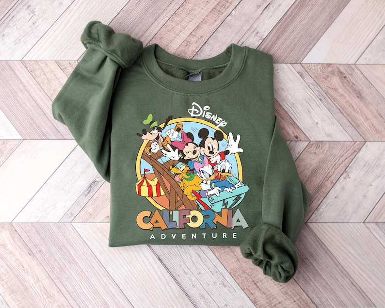 May include: An olive green sweatshirt featuring a colorful graphic of Disney characters on a roller coaster. The graphic includes Mickey Mouse, Minnie Mouse, Donald Duck, and Goofy. The words "Disney California Adventure" are printed below the graphic.