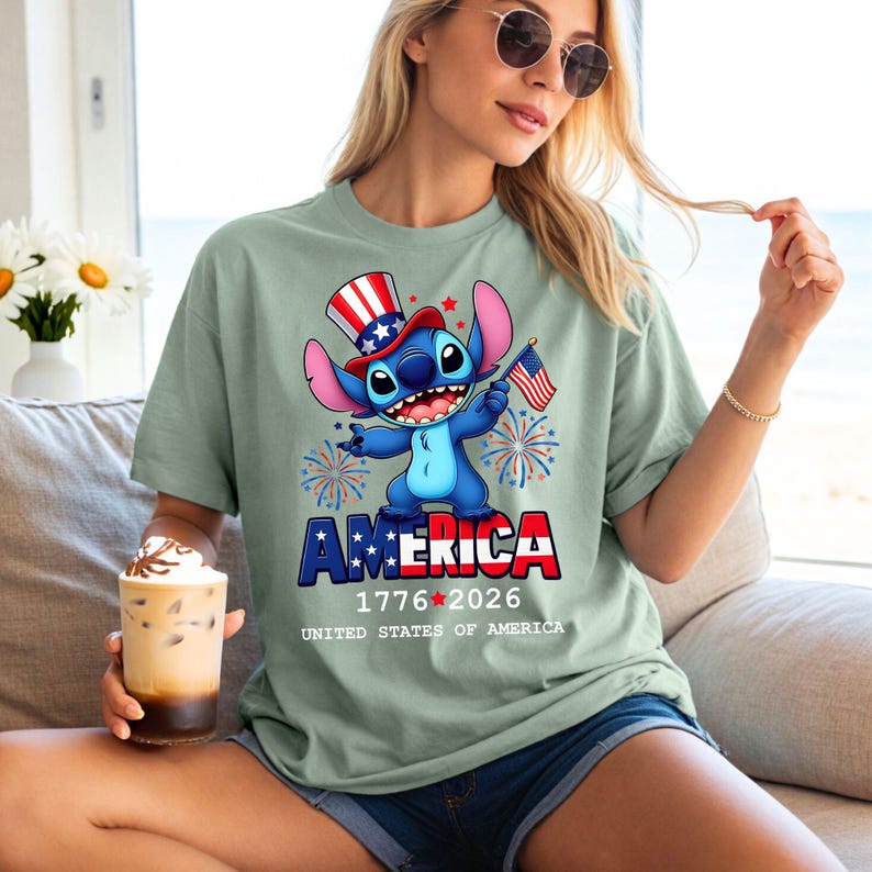 Puede incluir: Camiseta verde salvia con un personaje de dibujos animados que lleva un sombrero patri&oacute;tico y sostiene una peque&ntilde;a bandera estadounidense. La palabra "AMERICA" se muestra en rojo, blanco y azul, con los a&ntilde;os "1776-2026" debajo. Tambi&eacute;n se ve el texto "UNITED STATES OF AMERICA".