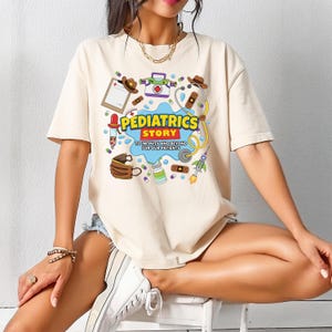 May include: A beige short-sleeve t-shirt featuring a colorful graphic print with medical-themed illustrations, including a stethoscope, bandages, and a clipboard. The text reads PEDIATRICS STORY TO INFINITY AND BEYOND FOR OUR PATIENTS.