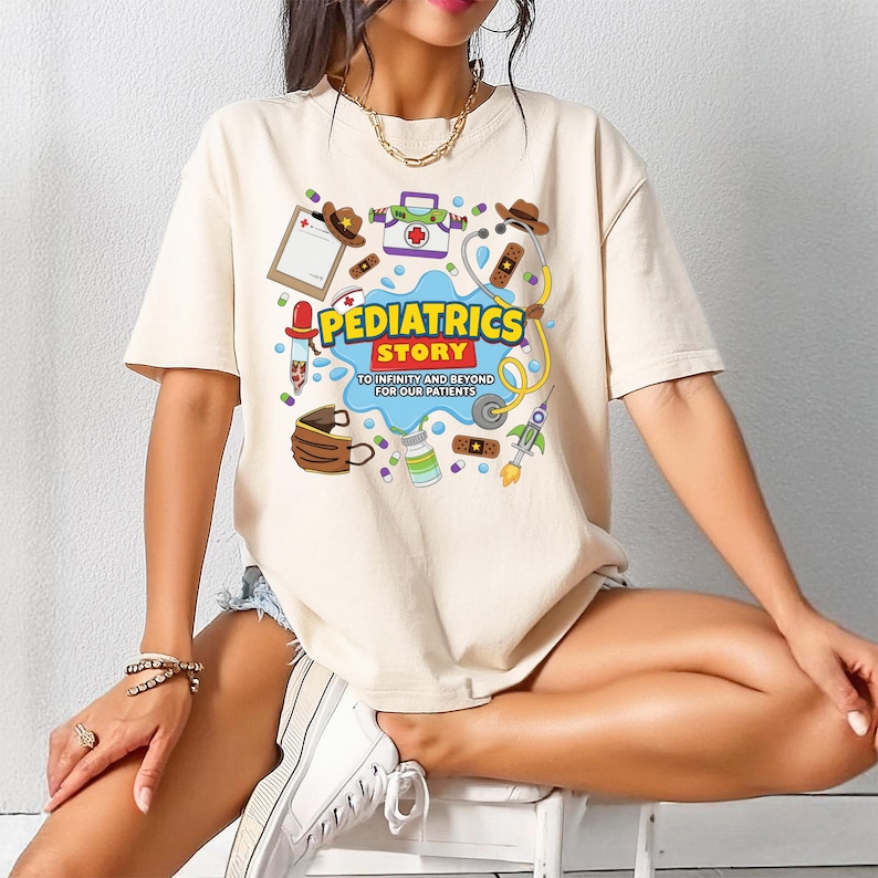 May include: A beige short-sleeve t-shirt featuring a colorful graphic print with medical-themed illustrations, including a stethoscope, bandages, and a clipboard. The text reads PEDIATRICS STORY TO INFINITY AND BEYOND FOR OUR PATIENTS.