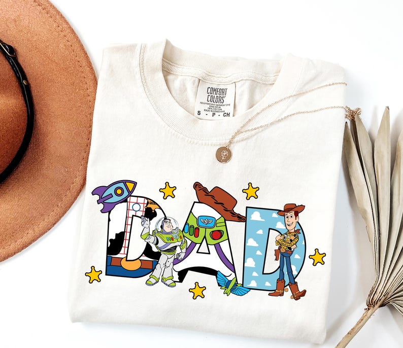 K&ouml;nnte beinhalten: Ein cremefarbenes T-Shirt mit dem Wort DAD in gro&szlig;en Buchstaben. Das D ist mit einer Rakete und Buzz Lightyear verziert, das A zeigt einen Cowboyhut und einen Raumanzug, und das D enth&auml;lt Woody. Gelbe Sterne umgeben den Schriftzug.