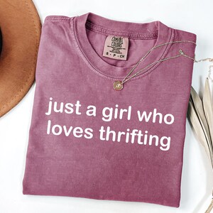 May include: A mauve-colored t-shirt with the text "just a girl who loves thrifting" in white. A gold necklace with a pendant rests on the shirt. A brown hat and dried plant are in the background.