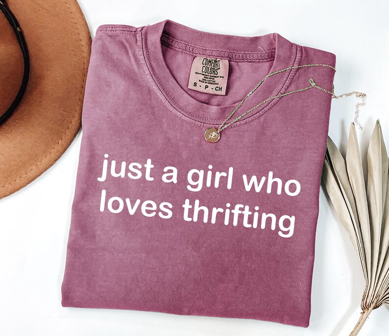 May include: A mauve-colored t-shirt with the text "just a girl who loves thrifting" in white. A gold necklace with a pendant rests on the shirt. A brown hat and dried plant are in the background.