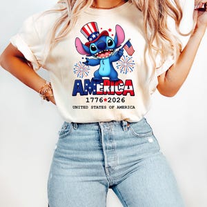 Puede incluir: Una camiseta color crema con un personaje de dibujos animados con un sombrero de copa patri&oacute;tico y una peque&ntilde;a bandera estadounidense. La palabra "AMERICA" se muestra en rojo y azul, con las fechas "1776-2026" debajo. Tambi&eacute;n se ve el texto "UNITED STATES OF AMERICA".