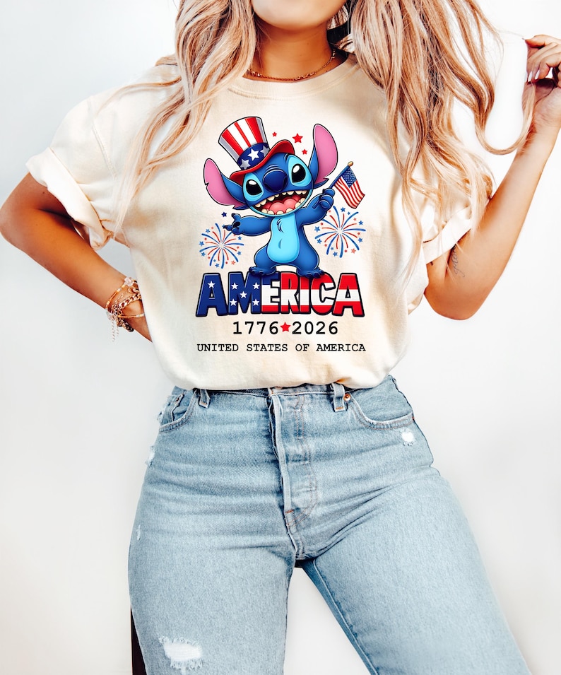 Puede incluir: Una camiseta color crema con un personaje de dibujos animados con un sombrero de copa patri&oacute;tico y una peque&ntilde;a bandera estadounidense. La palabra "AMERICA" se muestra en rojo y azul, con las fechas "1776-2026" debajo. Tambi&eacute;n se ve el texto "UNITED STATES OF AMERICA".