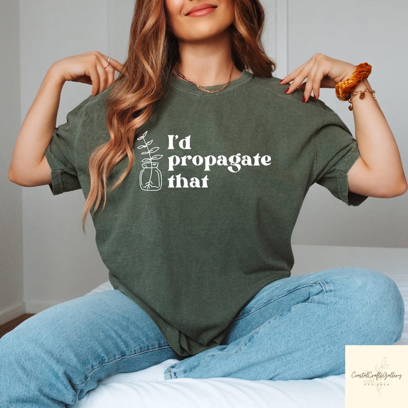 May include: Olive green t-shirt with the text "I'd propagate that" in white. The shirt features a line drawing of a plant in a jar. The model is wearing blue jeans.