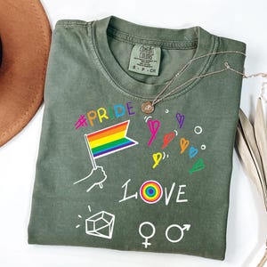 May include: An olive green short-sleeve t-shirt featuring a graphic print with the text #PRIDE and LOVE, a rainbow flag, hand-drawn hearts, a geometric shape, and gender symbols. The shirt has a Comfort Colors brand label at the inner neckline.
