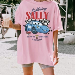 May include: A light pink t-shirt featuring a graphic of a blue cartoon car with the text "Lightning Sally". The design includes checkered flags and the words "Piston Cup Racing Series 5 Time Champ".