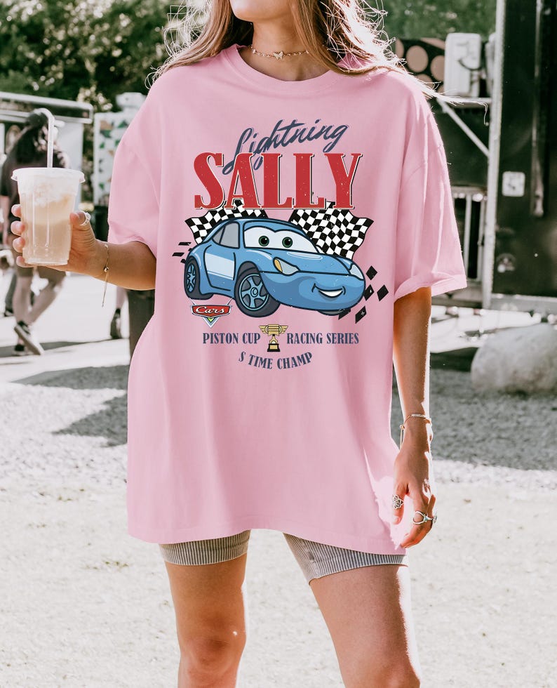 May include: A light pink t-shirt featuring a graphic of a blue cartoon car with the text "Lightning Sally". The design includes checkered flags and the words "Piston Cup Racing Series 5 Time Champ".