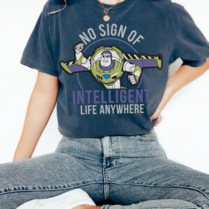 May include: A navy blue t-shirt with a Buzz Lightyear graphic and the text "NO SIGN OF INTELLIGENT LIFE ANYWHERE". The design incorporates green, purple, and white. This is a casual wear item.
