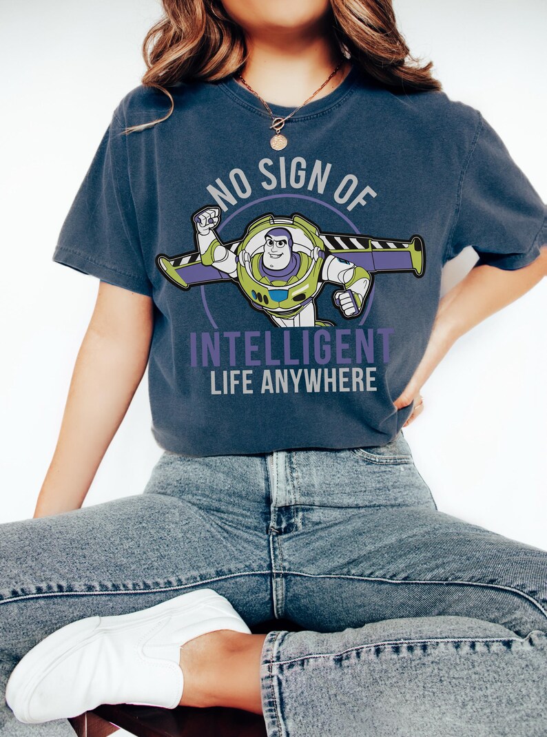 May include: A navy blue t-shirt with a Buzz Lightyear graphic and the text "NO SIGN OF INTELLIGENT LIFE ANYWHERE". The design incorporates green, purple, and white. This is a casual wear item.
