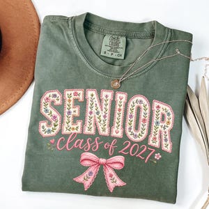 以下が含まれることがあります： オリーブグリーンの半袖Tシャツ。花柄で埋め尽くされた大きなクリーム色の文字で「SENIOR」と書かれ、その下にピンクの筆記体で「class of 2027」とプリントされています。Tシャツの下部には、花のアクセントが付いたピンクのリボンが描かれています。