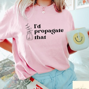 May include: A light pink t-shirt with the text "I'd propagate that" and a plant illustration. A white mug with a yellow smiley face is held in the hand. The shirt is paired with light blue shorts.