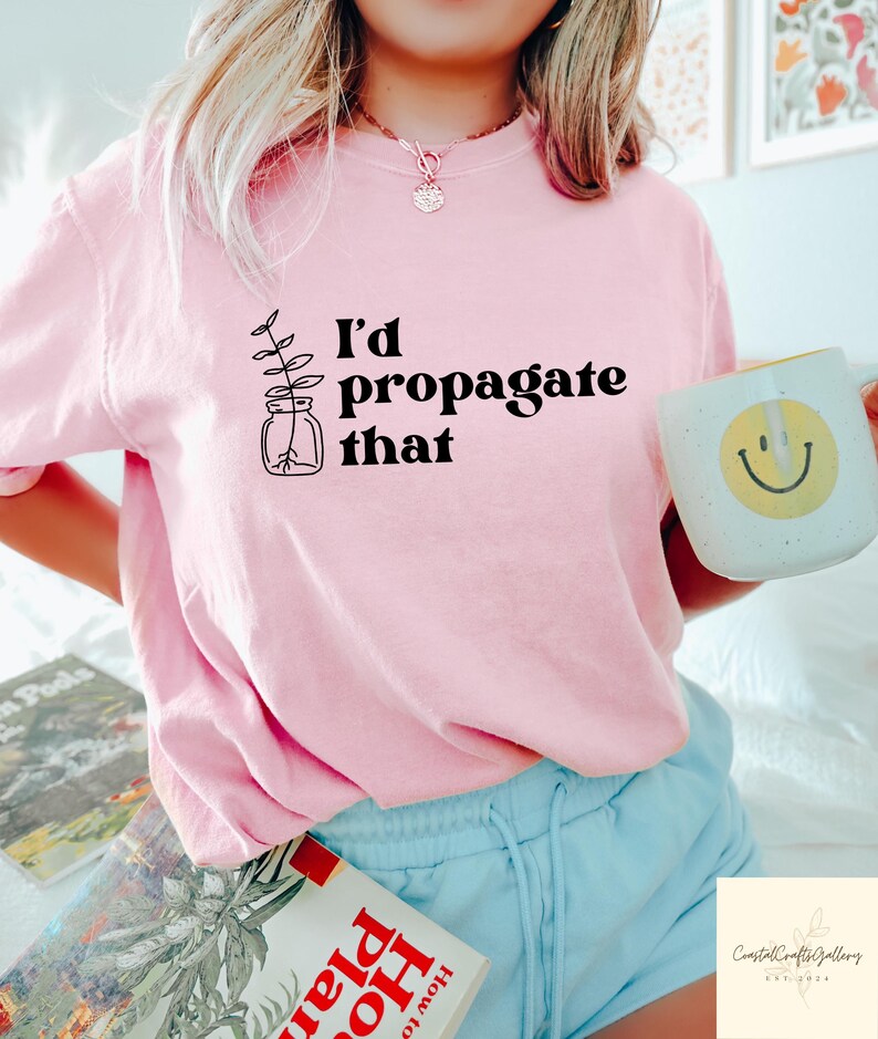 May include: A light pink t-shirt with the text "I'd propagate that" and a plant illustration. A white mug with a yellow smiley face is held in the hand. The shirt is paired with light blue shorts.