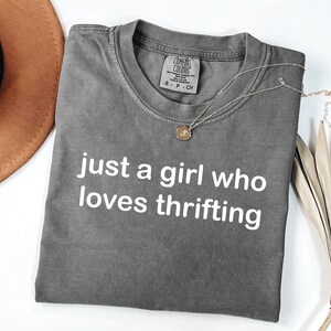 May include: A gray t-shirt with the text "just a girl who loves thrifting" in white. A gold necklace with a pendant rests on the shirt. A brown hat and dried palm leaves are also in the image.
