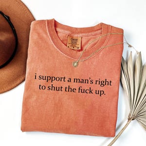 May include: Coral-colored Comfort Colors t-shirt with the text "i support a man's right to shut the fuck up." A gold necklace with a pendant and a brown hat are also visible. The shirt is folded on a white surface.