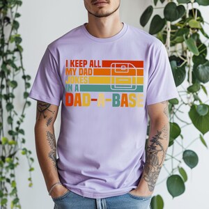 May include: A light purple t-shirt with the text "I KEEP ALL MY DAD JOKES IN A DAD-A-BASE" in a retro color scheme. The design includes a graphic of a floppy disk. The shirt is a casual wear item.