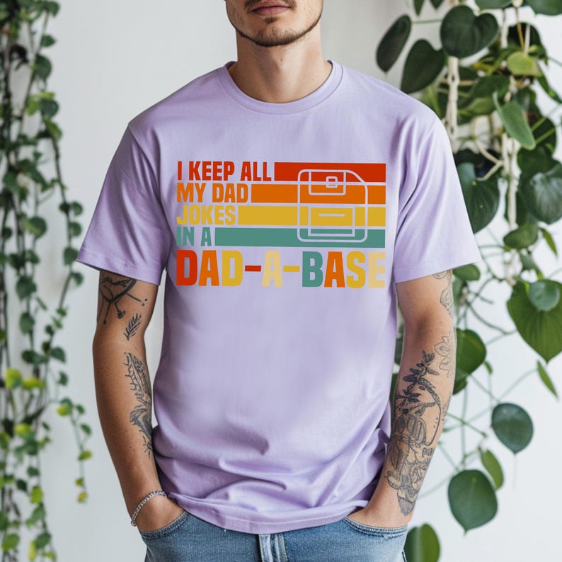 May include: A light purple t-shirt with the text "I KEEP ALL MY DAD JOKES IN A DAD-A-BASE" in a retro color scheme. The design includes a graphic of a floppy disk. The shirt is a casual wear item.