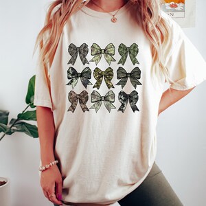 May include: Cream-colored t-shirt featuring a grid of nine bow designs in various camouflage patterns. The bows are in shades of green, gray, and brown. The shirt is worn by a person.