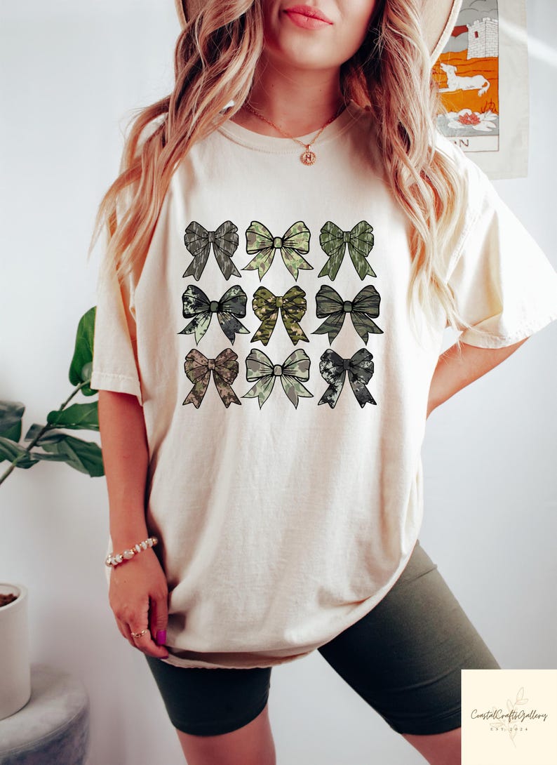 May include: Cream-colored t-shirt featuring a grid of nine bow designs in various camouflage patterns. The bows are in shades of green, gray, and brown. The shirt is worn by a person.
