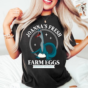 Puede incluir: Camiseta negra con una ilustraci&oacute;n de dibujos animados de un dinosaurio azul con una lengua roja. El texto dice "JOANNA'S FRESH FARM EGGS" en un dise&ntilde;o circular, con "DEFINITELY NOT STOLEN" debajo. La camiseta es oscura.