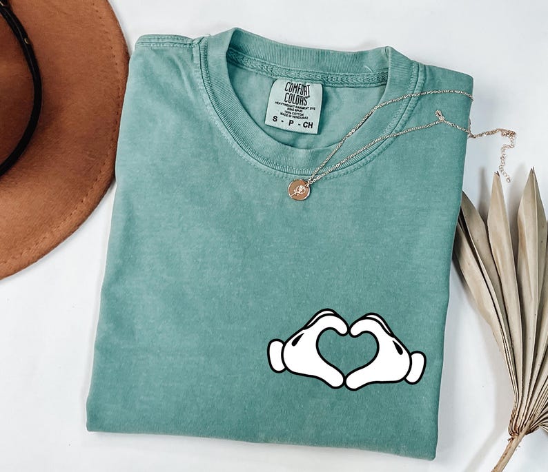 May include: A teal Comfort Colors t-shirt with a graphic of two white cartoon hands forming a heart shape. A gold necklace with a small pendant rests on the shirt. A brown hat and dried plant are in the background.