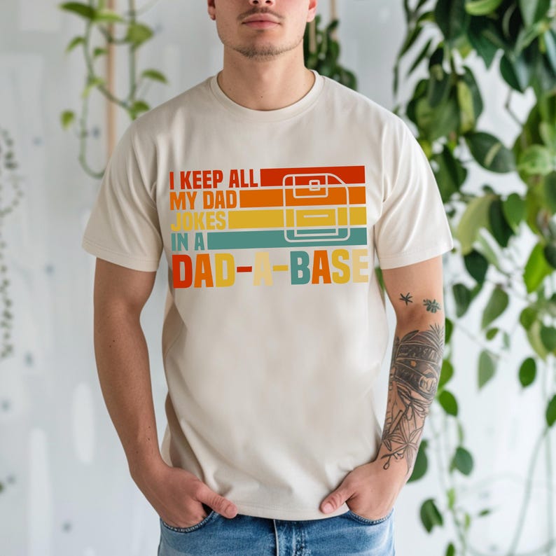 May include: A cream-colored t-shirt with the text "I KEEP ALL MY DAD JOKES IN A DAD-A-BASE" in a retro color scheme. The design includes a floppy disk graphic. The shirt is short-sleeved.