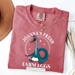 Puede incluir: Una camiseta de color rosa rojizo con un dibujo animado de un dinosaurio. La camiseta lleva el texto "Joanna's Fresh Farm Eggs" y "Definitely Not Stolen". Un collar de oro est&aacute; drapeado sobre la camiseta.