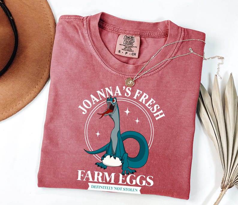 Puede incluir: Una camiseta de color rosa rojizo con un dibujo animado de un dinosaurio. La camiseta lleva el texto "Joanna's Fresh Farm Eggs" y "Definitely Not Stolen". Un collar de oro est&aacute; drapeado sobre la camiseta.