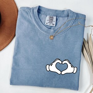May include: A blue short-sleeve t-shirt with a heart-shaped hand graphic. The shirt has a crew neck and a small tag on the collar. A gold necklace with a pendant is draped across the shirt.