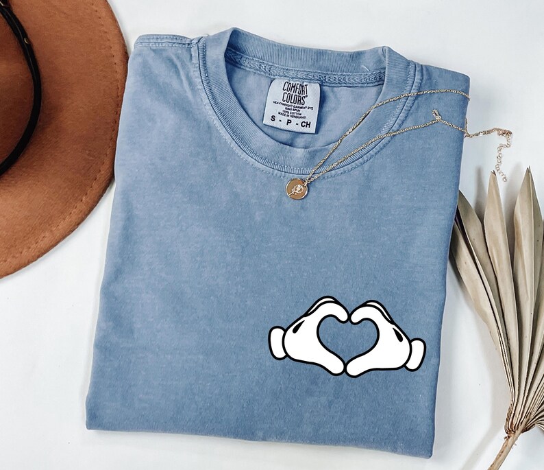 May include: A blue short-sleeve t-shirt with a heart-shaped hand graphic. The shirt has a crew neck and a small tag on the collar. A gold necklace with a pendant is draped across the shirt.