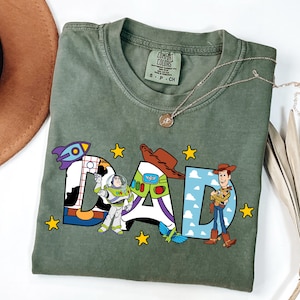 K&ouml;nnte beinhalten: Ein olivgr&uuml;nes Kurzarm-T-Shirt mit dem Wort DAD in gro&szlig;en Buchstaben. Die Buchstaben sind mit Toy Story-Charakteren verziert, darunter Buzz Lightyear, Woody, eine Rakete und gelbe Sterne. Das T-Shirt hat ein Comfort Colors Markenetikett.