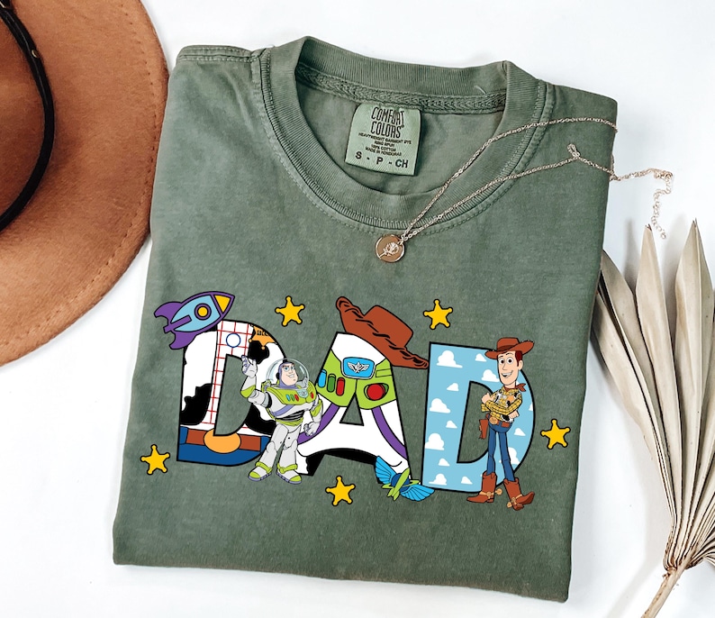 K&ouml;nnte beinhalten: Ein olivgr&uuml;nes Kurzarm-T-Shirt mit dem Wort DAD in gro&szlig;en Buchstaben. Die Buchstaben sind mit Toy Story-Charakteren verziert, darunter Buzz Lightyear, Woody, eine Rakete und gelbe Sterne. Das T-Shirt hat ein Comfort Colors Markenetikett.