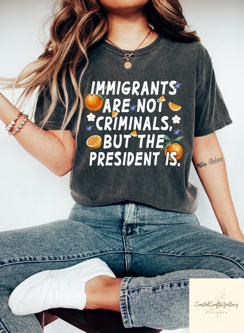 May include: Dark gray t-shirt with the text "IMMIGRANTS ARE NOT CRIMINALS, BUT THE PRESIDENT IS." Oranges and flowers are used as design elements. The person is wearing blue jeans and white sneakers.
