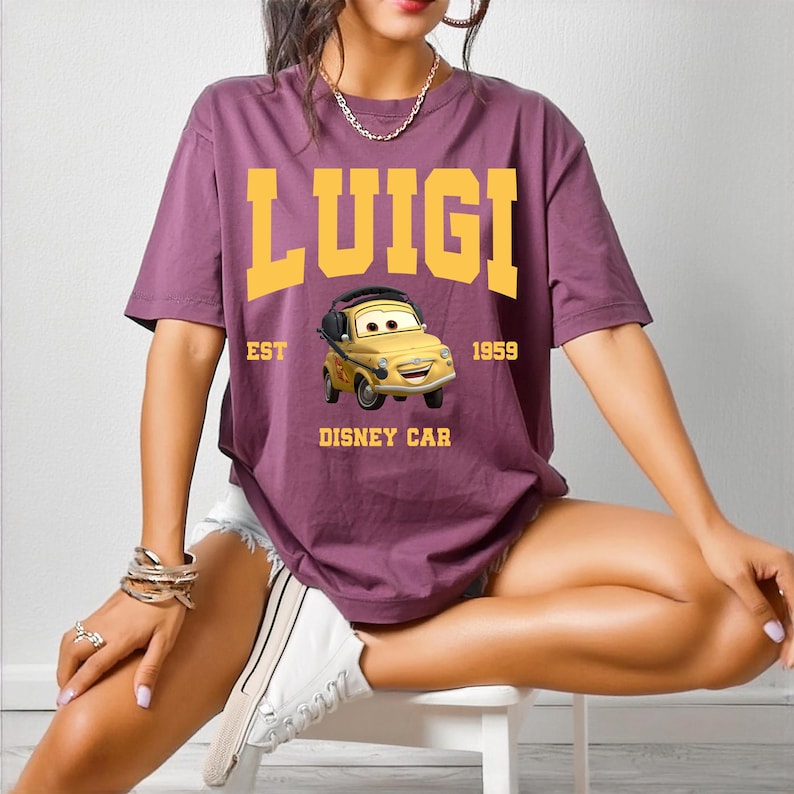 May include: A purple t-shirt featuring the word "LUIGI" in yellow block letters above a cartoon car graphic. The graphic includes the text "EST 1959 DISNEY CAR". The shirt is short-sleeved.