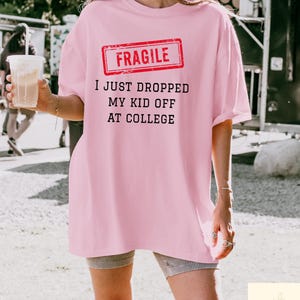 May include: A light pink t-shirt with the text "FRAGILE" in a red rectangle, and the phrase "I JUST DROPPED MY KID OFF AT COLLEGE" in black. The shirt is a loose fit.