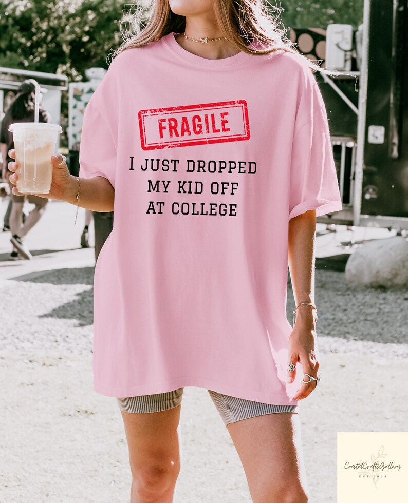 May include: A light pink t-shirt with the text "FRAGILE" in a red rectangle, and the phrase "I JUST DROPPED MY KID OFF AT COLLEGE" in black. The shirt is a loose fit.