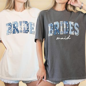 Puede incluir: Dos camisetas holgadas, una crema y otra gris carb&oacute;n. La camiseta crema dice "BRIDE" en letras azules de patchwork. La camiseta gris dice "BRIDESMAID" en letras azules de patchwork con "maid" en cursiva debajo. Ambas camisetas tienen mangas cortas.