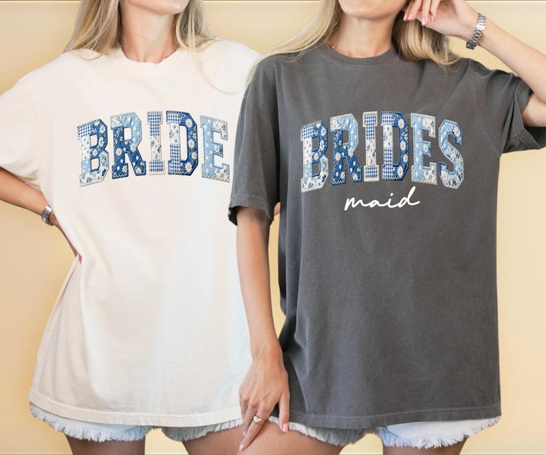 Puede incluir: Dos camisetas holgadas, una crema y otra gris carb&oacute;n. La camiseta crema dice "BRIDE" en letras azules de patchwork. La camiseta gris dice "BRIDESMAID" en letras azules de patchwork con "maid" en cursiva debajo. Ambas camisetas tienen mangas cortas.