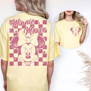 Puede incluir: Camiseta amarilla clara con un gr&aacute;fico de Minnie Mouse. La parte trasera presenta un gran dise&ntilde;o de Minnie Mouse sobre un fondo a cuadros rosa, con las palabras "Minnie Mouse". La parte delantera tiene un gr&aacute;fico de Minnie Mouse m&aacute;s peque&ntilde;o.