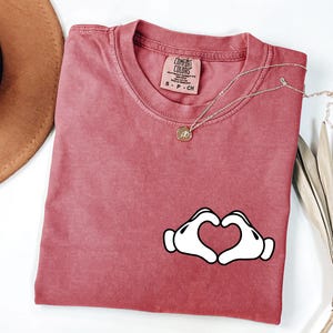 May include: A dusty rose Comfort Colors t-shirt with a white heart-shaped hand graphic. A gold necklace with a small pendant rests on the shirt. The shirt has a crew neck and a small tag.