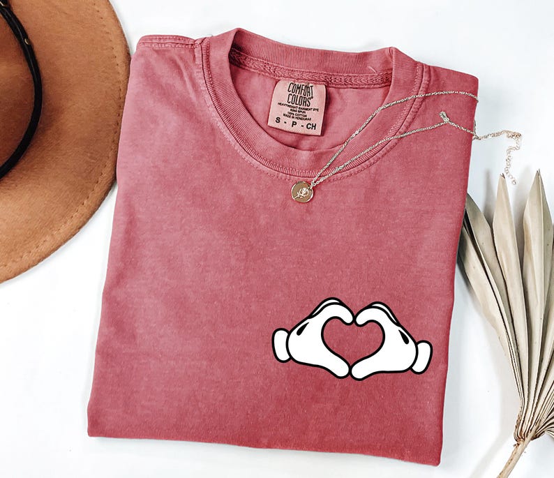 May include: A dusty rose Comfort Colors t-shirt with a white heart-shaped hand graphic. A gold necklace with a small pendant rests on the shirt. The shirt has a crew neck and a small tag.