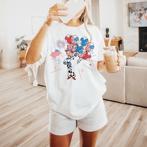 May include: White t-shirt featuring a cartoon illustration of a character holding balloons with patriotic designs. The design includes red, white, and blue elements, including fireworks and an American flag. The shirt is paired with gray shorts.