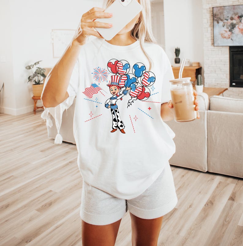 May include: White t-shirt featuring a cartoon illustration of a character holding balloons with patriotic designs. The design includes red, white, and blue elements, including fireworks and an American flag. The shirt is paired with gray shorts.