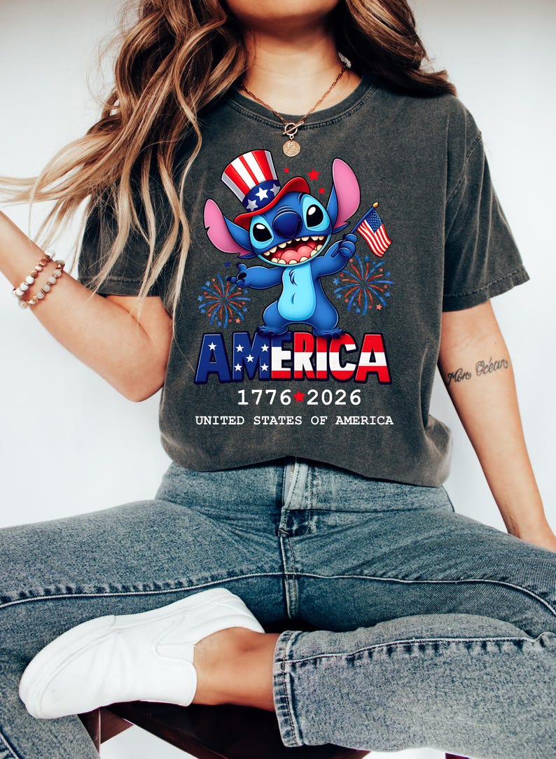 Puede incluir: Camiseta gris oscuro con un personaje de dibujos animados que lleva un sombrero patri&oacute;tico y sostiene una peque&ntilde;a bandera estadounidense. La palabra "AMERICA" est&aacute; en rojo y azul, con las fechas 1776-2026 debajo. La camiseta se combina con vaqueros azules.