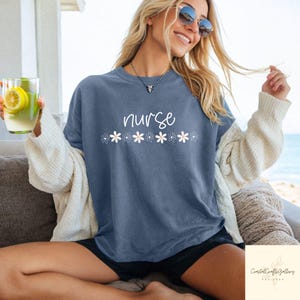 May include: A blue t-shirt with the word "nurse" in white script, and a row of white daisy graphics. The shirt is a relaxed fit. The image also shows a person wearing sunglasses and holding a glass of lemonade.